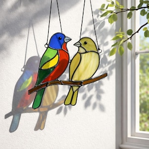 Puede incluir: Una pieza de arte en vitral con dos pájaros coloridos posados en una rama marrón. Uno tiene tonos azules, verdes, amarillos y rojos, el otro es amarillo. Los pájaros están suspendidos por cables negros, proyectando una sombra colorida.
