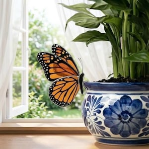 May include: A decorative monarch butterfly with orange and black wings rests on a blue and white floral patterned ceramic pot. The pot holds a green plant, set near a window with a view of greenery and white curtains.