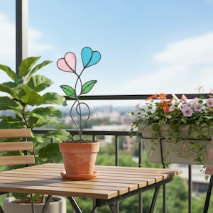 May include: A decorative garden stake with heart-shaped pink and blue glass flowers and green leaves. The stake is in a terracotta pot on a wooden table. The background shows a cityscape.