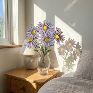 May include: A clear glass vase holding a bouquet of daisy-like flowers with purple petals and yellow centers. The flowers are on a wooden nightstand next to a lamp. The sunlight casts shadows on the wall.