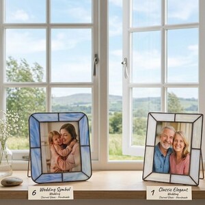 K&ouml;nnte beinhalten: Zwei Glasmalerei-Fotorahmen mit schwarzen Metallr&auml;ndern stehen auf einer Holzoberfl&auml;che vor einem Fenster. Ein Rahmen ist blau, der andere wei&szlig;. Jeder Rahmen enth&auml;lt ein Foto eines Paares. Der Text auf der Unterseite der Rahmen lautet "Wedding Symbol" und "Classic Elegant".