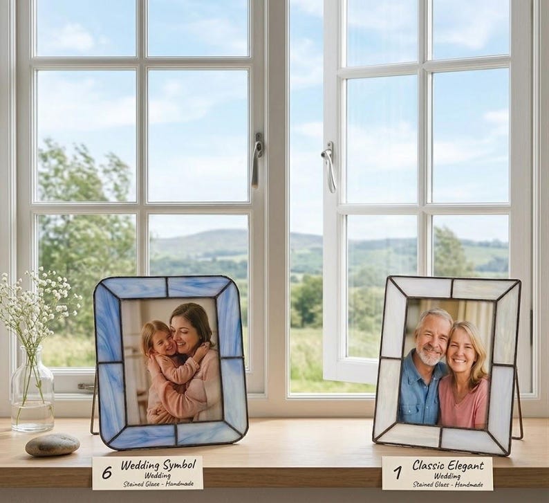 K&ouml;nnte beinhalten: Zwei Glasmalerei-Fotorahmen mit schwarzen Metallr&auml;ndern stehen auf einer Holzoberfl&auml;che vor einem Fenster. Ein Rahmen ist blau, der andere wei&szlig;. Jeder Rahmen enth&auml;lt ein Foto eines Paares. Der Text auf der Unterseite der Rahmen lautet "Wedding Symbol" und "Classic Elegant".