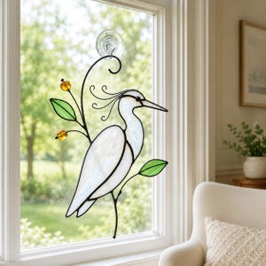 May include: A stained glass suncatcher featuring a white heron with black outlines, perched on a branch with green leaves and two amber-colored glass beads. The piece is suspended by a clear suction cup attached to a window pane.