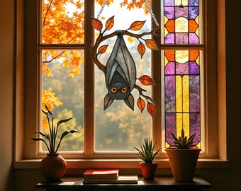 May include: A stained glass bat decoration hanging in front of a window. The bat is grey with orange eyes and is suspended upside down from a tree branch with orange leaves. The window features a colourful stained glass panel and a view of autumn foliage.