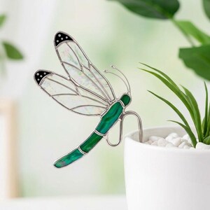 Può includere: Un ornamento a forma di libellula in vetro colorato con ali bianche iridescenti dalle punte nere e un corpo verde solido. La libellula è realizzata con sottili linee di saldatura in metallo e include un gambo in filo metallico curvo.