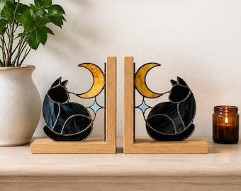 Puede incluir: Un par de sujetalibros de madera en forma de L con inserciones de vidrieras. Cada inserción representa un gato negro sentado bajo una luna creciente amarilla y una pequeña estrella azul claro de cuatro puntas, unidas por líneas de soldadura.