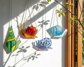 May include: Four colorful stained glass snail suncatchers hanging from chains. The snails feature green, orange, red, blue, and purple shells with various patterns. The suncatchers are hanging in front of a white wall with shadows.