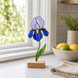 Könnte beinhalten: Eine Iris-Blumenskulptur aus Buntglas mit hellvioletten oberen Blütenblättern, dunkelblauen unteren Blütenblättern und einer gelben Mitte. Die Blume ist an einem dünnen schwarzen Metallstiel mit einem grünen Blatt auf einem Holzsockel befestigt.