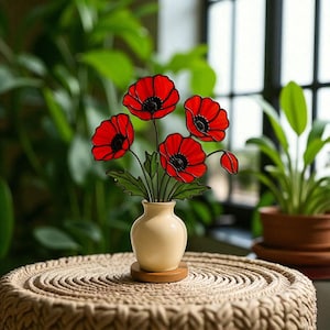 May include: A decorative arrangement of red poppy flowers with black centers and green leaves in a cream-colored vase. The vase sits on a small wooden base, placed on a woven table.