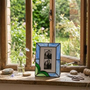 K&ouml;nnte beinhalten: Ein Glasmalerei-Fotorahmen mit einem blau-gr&uuml;nen Blattdesign. Der Rahmen enth&auml;lt zwei Schwarz-Wei&szlig;-Fotos. Der Rahmen steht auf einer Holzfensterbank.