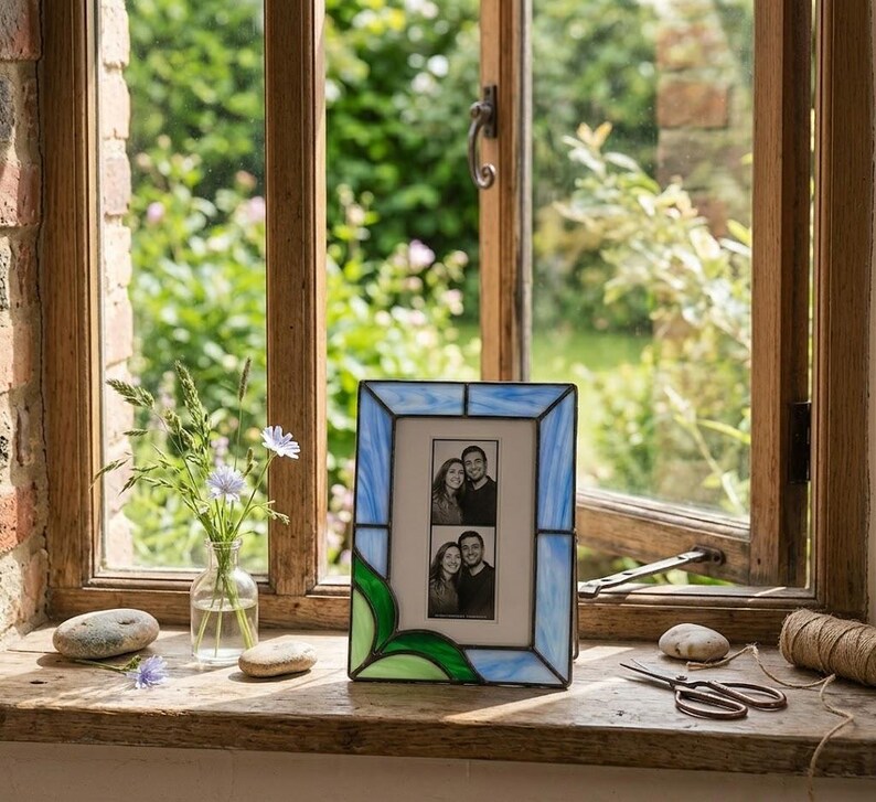 K&ouml;nnte beinhalten: Ein Glasmalerei-Fotorahmen mit einem blau-gr&uuml;nen Blattdesign. Der Rahmen enth&auml;lt zwei Schwarz-Wei&szlig;-Fotos. Der Rahmen steht auf einer Holzfensterbank.