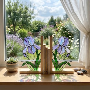 May include: Stained glass iris flower bookends in shades of purple, blue, and green, with wooden bases. The bookends are holding a stack of books on a wooden windowsill, with a small succulent plant in a ceramic pot to the left.
