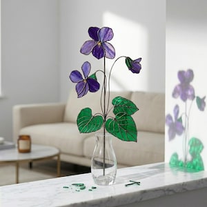 May include: A stained glass violet flower arrangement with purple petals and green leaves. The flowers are in a clear glass vase. The arrangement is on a white marble surface.