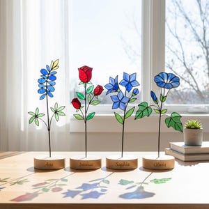 以下が含まれることがあります： 青と赤の色合いのステンドグラスの花の彫刻が4つ。それぞれ木製の台座に取り付けられています。台座にはAva、Jessica、Sophia、Oliviaの名前が刻まれています。花には、ルピナス、バラ、アサガオが含まれています。