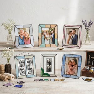 K&ouml;nnte beinhalten: Eine Sammlung von Glasmalerei-Fotorahmen in verschiedenen Formen und Gr&ouml;&szlig;en. Jeder Rahmen hat ein anderes Farbschema, darunter Blau, Rosa und Pastellt&ouml;ne. Die Rahmen enthalten Fotos von Menschen und werden auf einer Holzoberfl&auml;che pr&auml;sentiert.