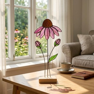 May include: A stained glass flower sculpture with a pink and brown flower head, two pink buds, and green leaves. The sculpture is mounted on a small wooden base. The sculpture is on a wooden table near a window.