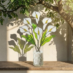 May include: A glass vase filled with white pebbles holds a bouquet of stained glass calla lilies. The lilies are white with yellow centers and green leaves. The vase sits on a wooden surface, with the shadow of the flowers cast on the wall behind.
