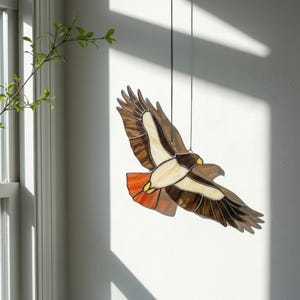 May include: A stained glass hawk in flight, suspended by black strings. The bird features brown, cream, and orange hues, with detailed wing and feather patterns. The artwork is displayed against a white wall, with natural light casting shadows.