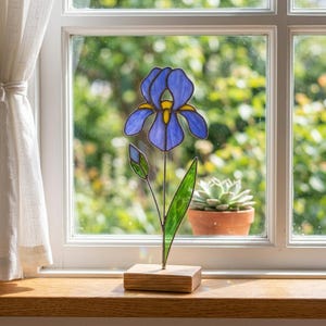 Può includere: Una scultura di fiore di iris in vetro colorato con petali blu e dettagli gialli, montata su una piccola base di legno. Il fiore ha uno stelo e una foglia verdi. La scultura è esposta su un davanzale.