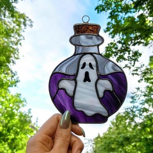 Könnte beinhalten: Glasmalerei-Dekoration in Form einer Zaubertrankflasche mit einem Geist darin. Die Flasche hat einen silbernen Rahmen, einen Korken und ist mit violettem und weißem Glas gefüllt. Der Geist ist weiß mit einem schwarzen Mund.