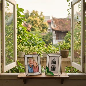 K&ouml;nnte beinhalten: Zwei dekorative Fotorahmen mit schwarzen Metallst&auml;ndern. Ein Rahmen enth&auml;lt ein Farbfoto eines Paares, der andere ein Schwarz-Wei&szlig;-Foto einer Familie. Die Rahmen stehen auf einem Holzregal vor einem offenen Fenster.