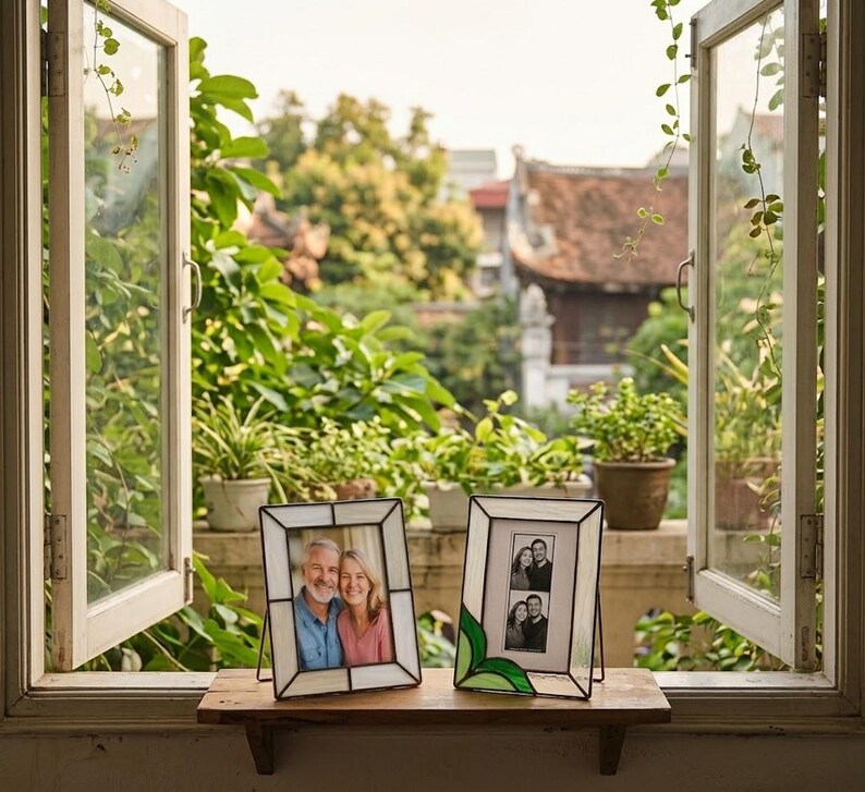 K&ouml;nnte beinhalten: Zwei dekorative Fotorahmen mit schwarzen Metallst&auml;ndern. Ein Rahmen enth&auml;lt ein Farbfoto eines Paares, der andere ein Schwarz-Wei&szlig;-Foto einer Familie. Die Rahmen stehen auf einem Holzregal vor einem offenen Fenster.