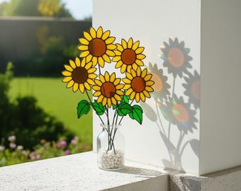 Puede incluir: Un jarrón de cristal lleno de guijarros blancos contiene un ramo de girasoles amarillos y marrones con hojas verdes. Los girasoles están frente a una pared blanca, con la luz del sol proyectando una sombra de las flores.