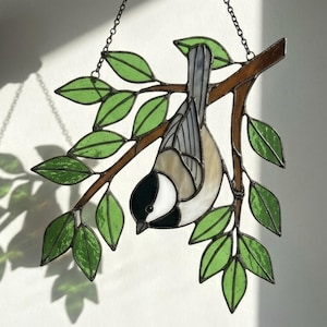 May include: Stained glass artwork of a chickadee bird hanging upside down from a tree branch. The bird has black, white, and tan coloring. The leaves are green, and the branch is brown. A metal chain is attached for hanging.