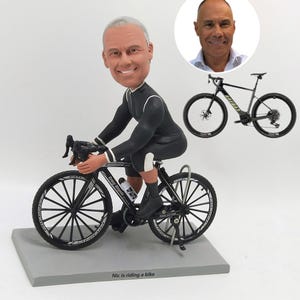 K&ouml;nnte beinhalten: Figur eines Radfahrers auf einem schwarz-wei&szlig;en Fahrrad. Der Radfahrer tr&auml;gt einen schwarz-wei&szlig;en Radanzug. Die Figur steht auf einem grauen Sockel mit dem Text "Nic is riding a bike". Im Hintergrund ein Bild eines Mannes und eines Fahrrads.