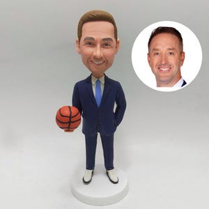 May include: A bobblehead figurine of a man in a navy blue suit, holding a basketball. The figurine has a smiling face and is on a white base. A smaller image of the man is in the background.