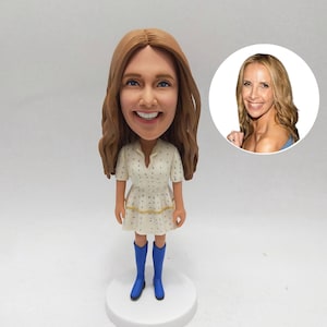 May include: A custom bobblehead figurine of a woman with long brown hair, wearing a white dress with blue polka dots and blue boots. A smaller image of the woman is in the background.