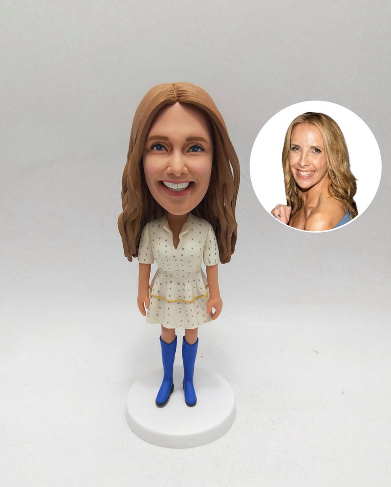 May include: A custom bobblehead figurine of a woman with long brown hair, wearing a white dress with blue polka dots and blue boots. A smaller image of the woman is in the background.
