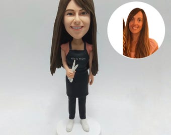 May include: A bobblehead figurine of a smiling woman with long brown hair, wearing a black apron with the text "Hair by Megan". She holds scissors and is dressed in black trousers and white shoes. A smaller image of the woman is in the background.