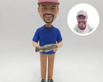 May include: A custom bobblehead figurine of a person wearing a blue shirt, tan pants, and a baseball cap, holding a fish. The figurine is on a white base, with a smaller image of the person in a circle.