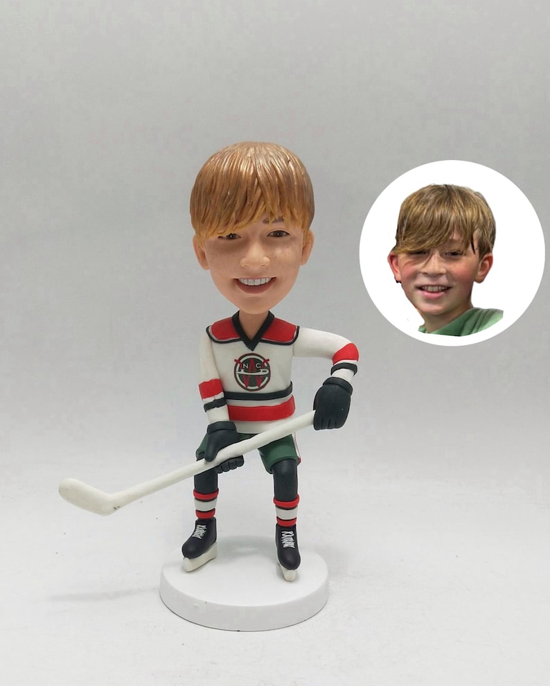 May include: A bobblehead figurine of a hockey player in a white, red, and black jersey, holding a hockey stick. The figurine has a smiling face and is on a white base. A smaller image of a boy is in the background.