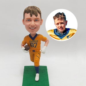 May include: A custom bobblehead figurine of a young boy wearing a yellow and blue football uniform with the number 87 on the front. The figurine is holding a football and is running. The figurine is on a green base.