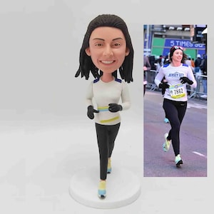 May include: Custom running figurine, a caricature of a woman in running attire. The figurine is wearing a white long-sleeve shirt with a yellow and blue stripe across the chest, black pants, and light blue running shoes.  The figurine is posed mid-stride. A photograph of a woman running in a race is shown next to the figurine. The woman in the photo wears a white shirt with 'Jersey City' and race number 'B 7862' printed on it.  The figurine is a personalized gift, perfect for runners or marathon participants.