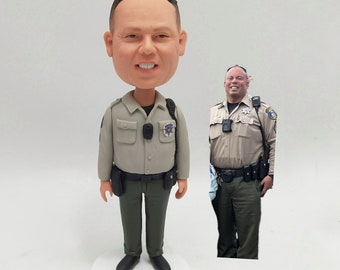 May include: A custom-made figurine of a person in a green and brown uniform with a badge. The figurine has a large head and a small body. The person is smiling and has a friendly expression.