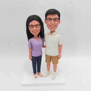 K&ouml;nnte beinhalten: Eine personalisierte Wackelkopf-Figur eines Paares. Die Frau tr&auml;gt ein lila Oberteil, blaue Jeans und eine Brille. Der Mann tr&auml;gt ein wei&szlig;es Poloshirt, khakifarbene Shorts und eine Brille. Beide Figuren l&auml;cheln und stehen auf einem wei&szlig;en Sockel.