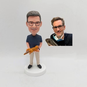 May include: A custom-made figurine of a person wearing glasses and a blue shirt, holding a small orange lizard. The figurine is standing on a white base.