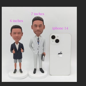 K&ouml;nnte beinhalten: Zwei Wackelkopf-Figuren von M&auml;nnern in Business-Kleidung, eine 15 cm und die andere 18 cm gro&szlig;, stehen auf wei&szlig;en, kreisf&ouml;rmigen Sockeln. Die gr&ouml;&szlig;ere Figur h&auml;lt ein schwarzes Buch. Ein iPhone 14 befindet sich neben den Figuren.