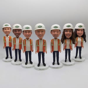 Könnte beinhalten: Eine Sammlung von Wackelkopf-Figuren von Personen, die weiße Schutzhelme mit dem Wort "PIRTLE" und orangefarbene Sicherheitswesten tragen. Jede Figur steht auf einem weißen Sockel mit einem aufgedruckten Namen. Die Figuren tragen weiße Hemden und dunkelblaue Hosen.