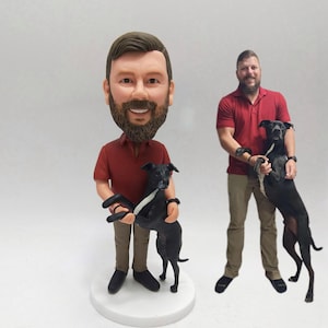 May include: A custom bobblehead figurine of a person in a red shirt and khaki pants, holding a black dog with a white stripe. The figurine is on a white base. Another person in a red shirt holds a black dog.