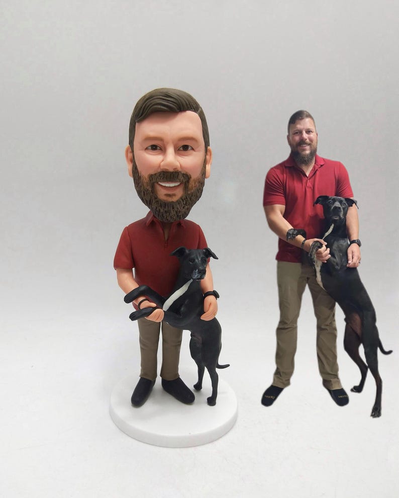 May include: A custom bobblehead figurine of a person in a red shirt and khaki pants, holding a black dog with a white stripe. The figurine is on a white base. Another person in a red shirt holds a black dog.