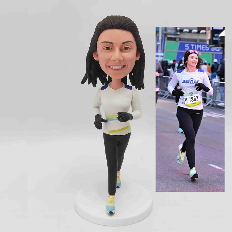May include: A running figurine of a woman in a white long-sleeve shirt with blue and yellow stripes, black leggings, and running shoes. The figurine is on a white base. A photo of a woman running a race is next to the figurine.