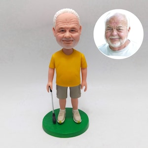 May include: A figurine of a man with white hair and a yellow shirt, khaki shorts, and beige sneakers, holding a golf club. The figurine stands on a green circular base. A smaller image of the man is in the background.