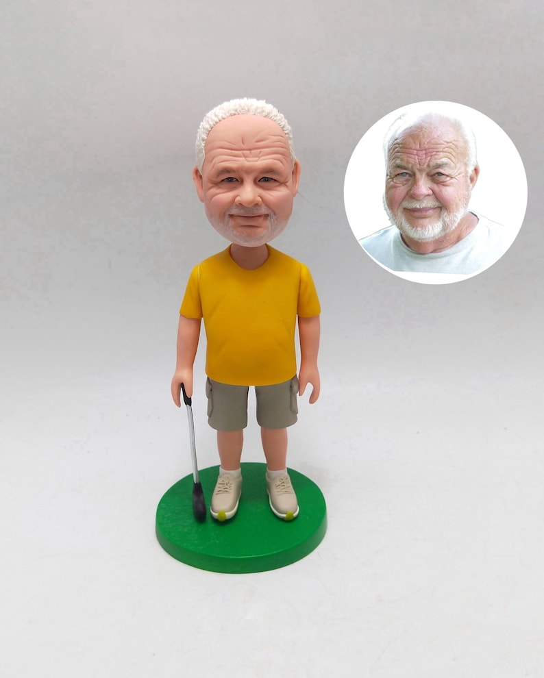 May include: A figurine of a man with white hair and a yellow shirt, khaki shorts, and beige sneakers, holding a golf club. The figurine stands on a green circular base. A smaller image of the man is in the background.
