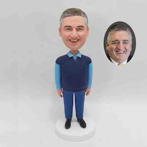 K&ouml;nnte beinhalten: Eine personalisierte Wackelkopf-Figur eines Mannes mit grauem Haar, der einen blauen Pullover &uuml;ber einem hellblauen Hemd, einer blauen Jeans und schwarzen Schuhen tr&auml;gt. Ein ovales Foto des Mannes befindet sich neben der Figur. Die Figur steht auf einer wei&szlig;en Basis.