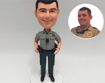 May include: A bobblehead figurine of a smiling person in a gray uniform, resembling a law enforcement officer. The figurine is standing on a white base, with a smaller circular image of the person in uniform. The uniform includes a badge and dark green pants.