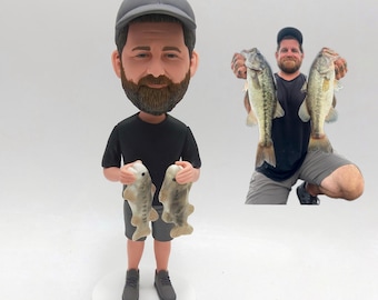 May include: A bobblehead figurine of a man wearing a black shirt, gray shorts, and a gray cap, holding two fish. A smaller image shows a man holding two fish. The figurine stands on a white base.